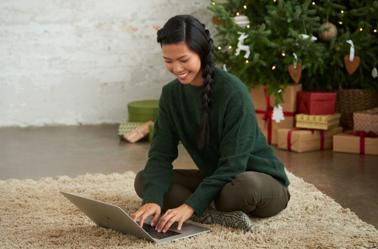 Henkilö istuu lattialla kannettavan tietokoneen edessä. Taustalla joulukuusi ja lahjoja.
