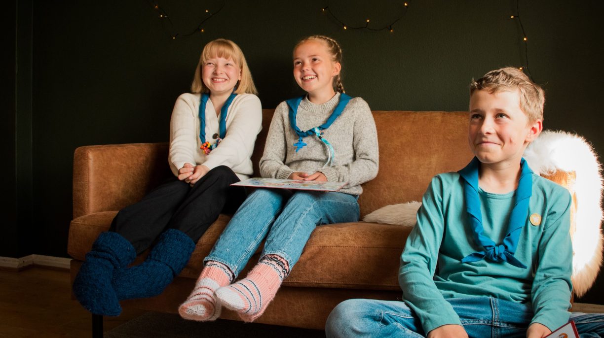 Kolme nuorta partiolaista hymyilemässä olohuoneessa. Kaksi istuu sohvalla ja kolmas lattialla.